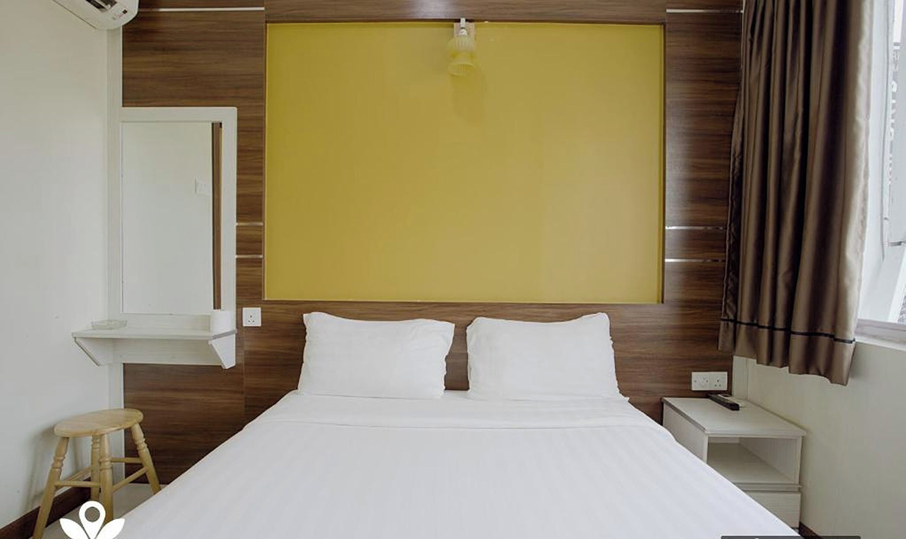 Une chambre d’hôtel comprenant un lit, une table de chevet, un petit tabouret, une étagère fixée au mur et une fenêtre avec des rideaux.