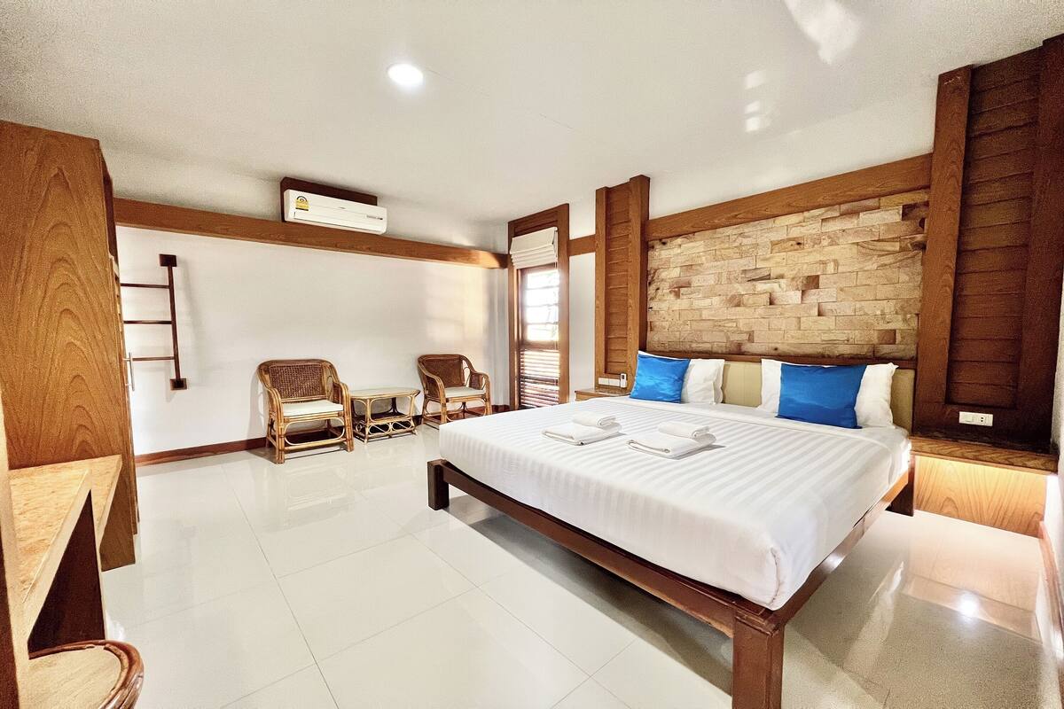 Habitación de hotel con una cama grande, cabecera de madera, dos sillones de mimbre, una mesita y un aire acondicionado instalado en la pared.