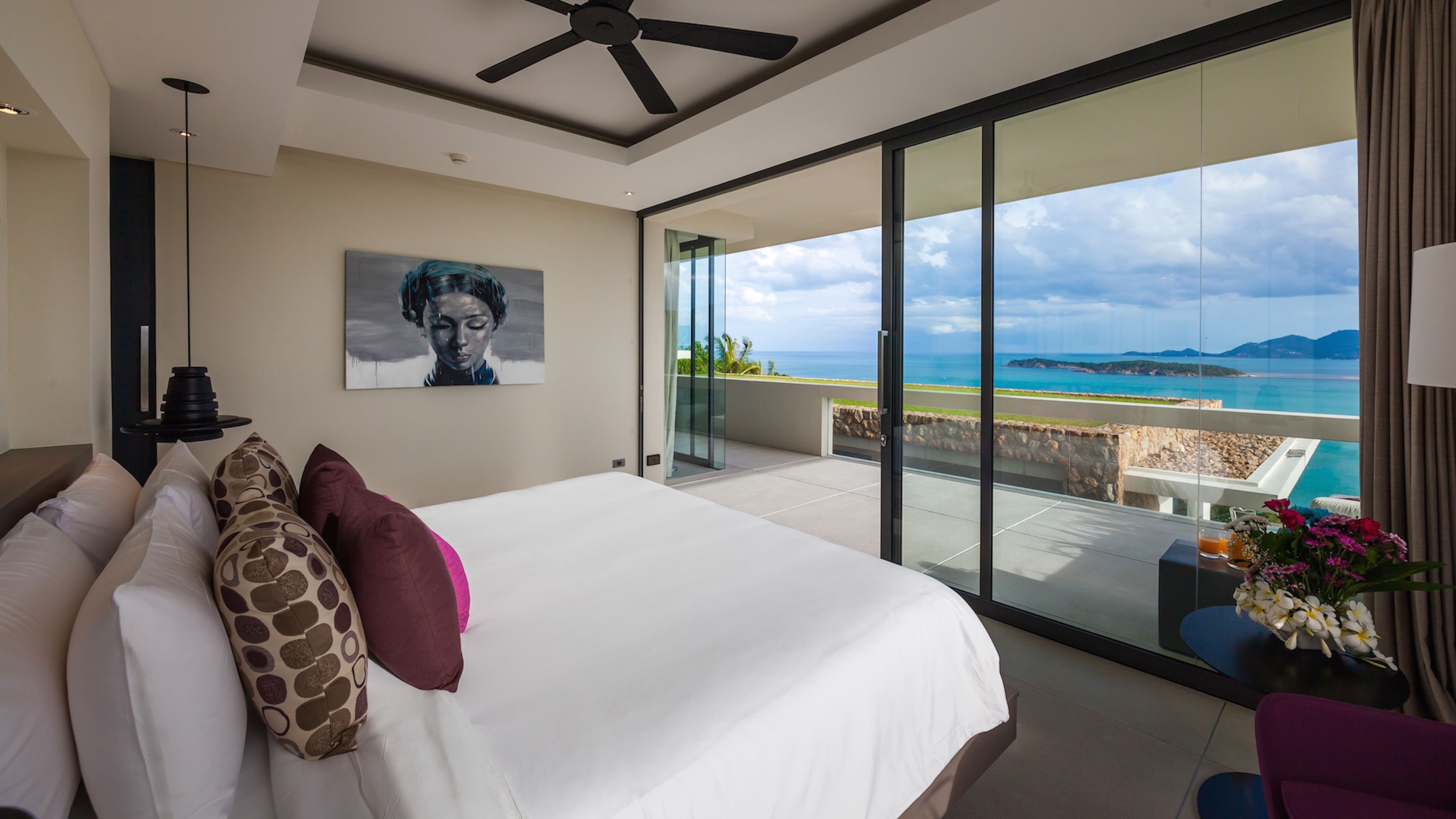 Une chambre d’hôtel moderne avec un grand lit, un ventilateur de plafond, un tableau et un balcon offrant une vue sur la mer.