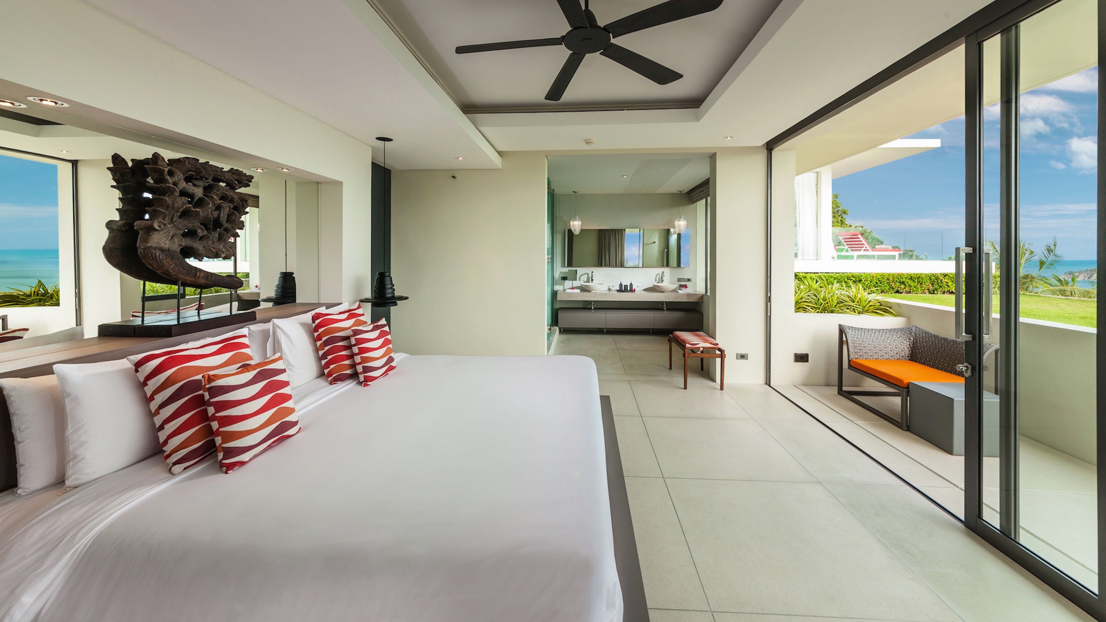 A modern hotel room with a large bed, a ceiling fan, a bathroom with a large mirror, and a balcony with a view of the ocean.
