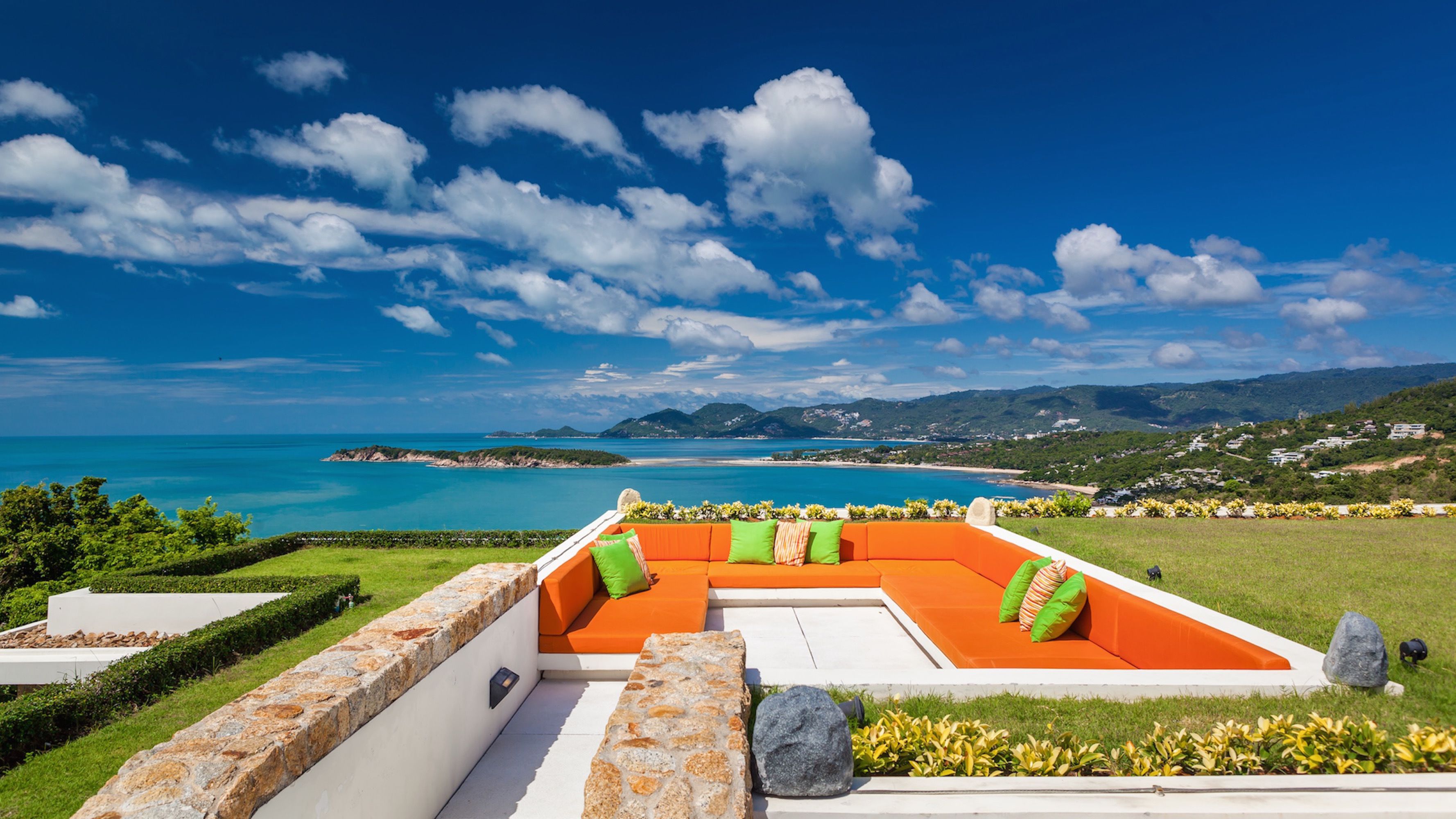An outdoor seating area with orange cushions and green pillows, overlooking a coastal landscape with clear blue skies and distant hills.