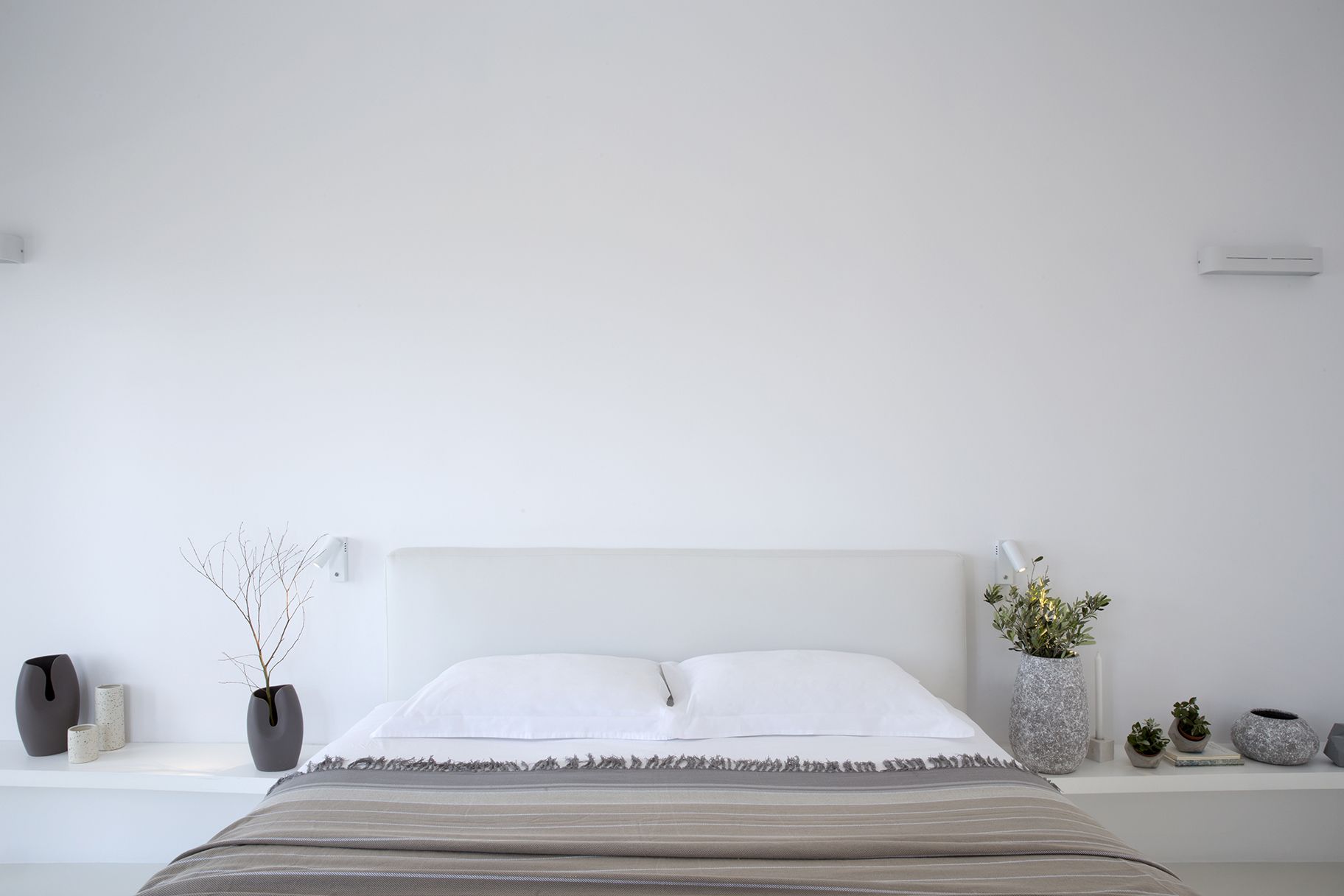A minimalist bedroom with a bed, white headboard, bedside tables with decorative items, and a plain wall.