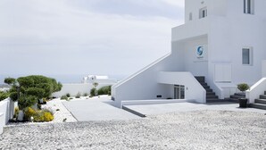 A modern white building with a logo, a staircase, and a view of the sea. - May Seven Santorini (Santorini)