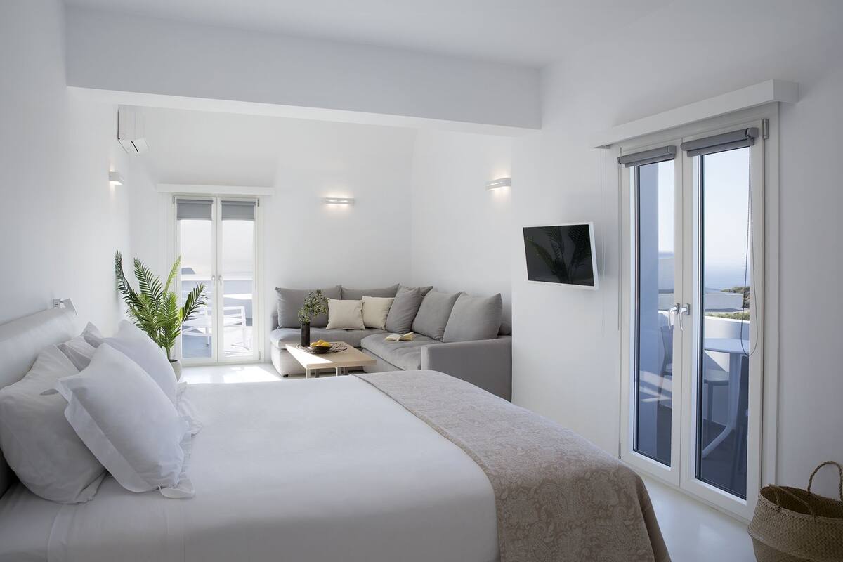A modern bedroom with a large bed, a sofa, a dining table, and a balcony with a view.