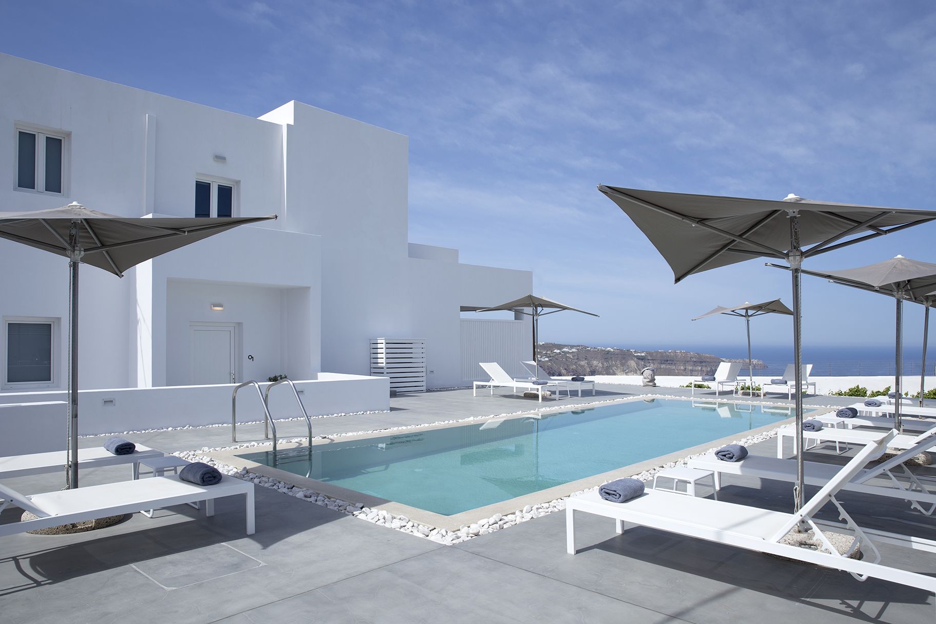 A rooftop pool area with white lounge chairs and umbrellas, a clear view of the ocean, and a modern white building.