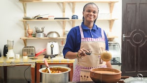 A person in a kitchen with wooden shelves, a pot on the counter, and a door in the background. - Nungwi House (Nungwi)