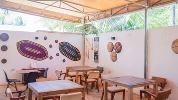 Un patio al aire libre con mesas y sillas de madera, una zona cubierta con techo metálico y detalles decorativos en las paredes.