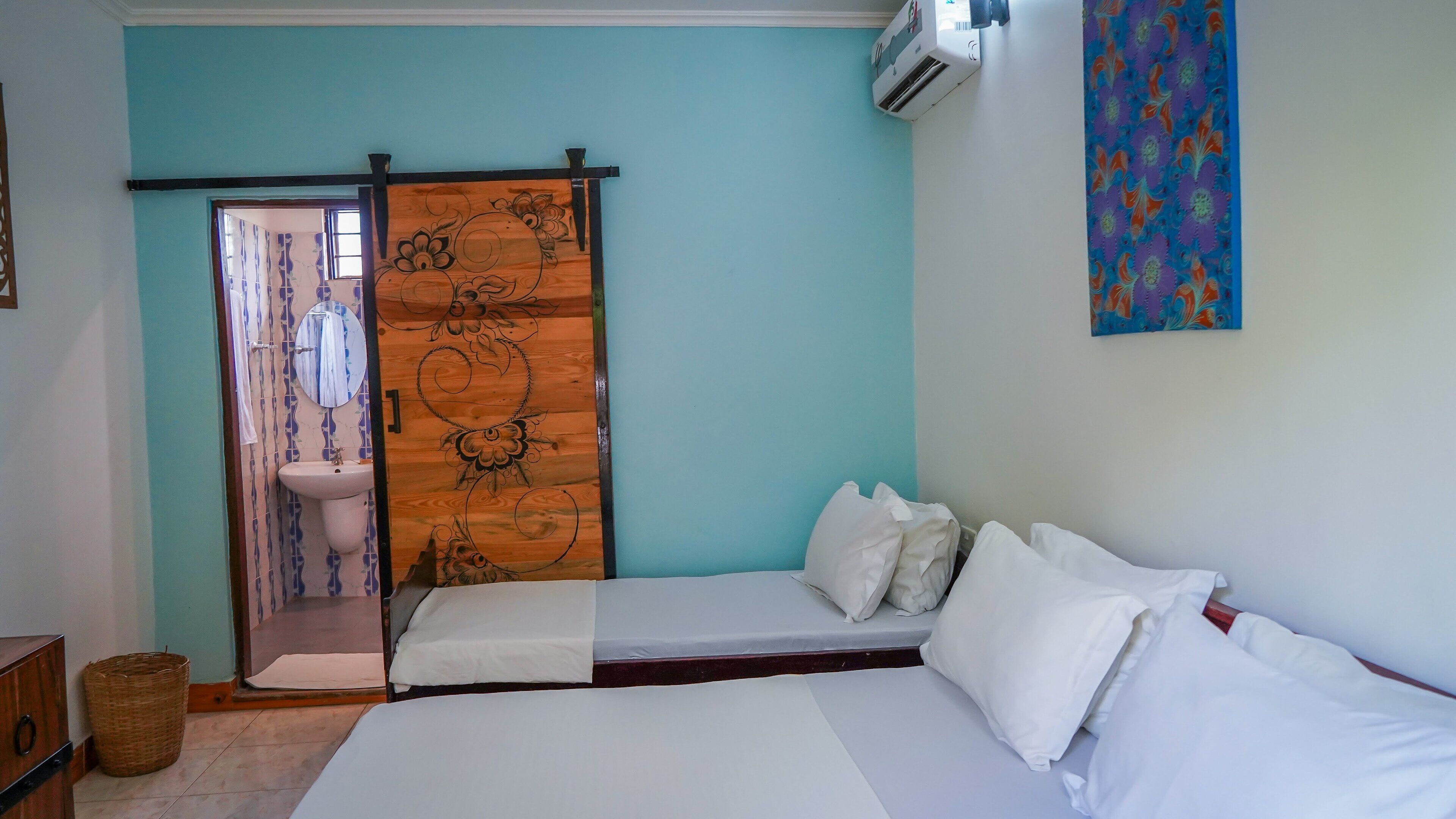 Un dormitorio con una cama, una puerta de madera con detalles florales, un lavamanos blanco y un aire acondicionado instalado en la pared.