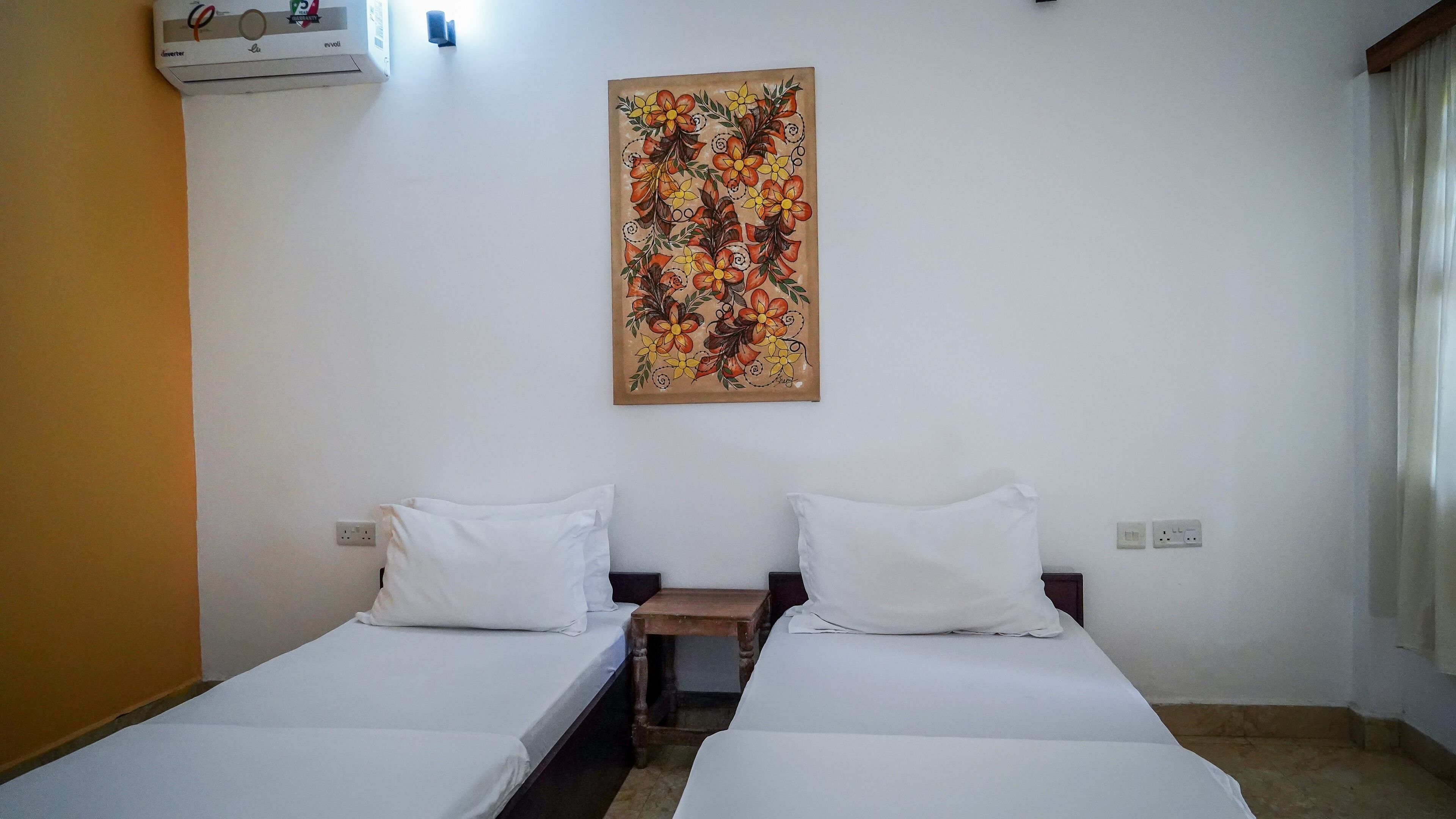 A hotel room with two single beds, a wall-mounted air conditioner, and a floral artwork above the beds.