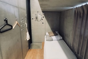 A small, compact room with a single bed, a wooden floor, a concrete wall, a hanging light fixture, and a coat rack with a hanger. - Kangaroo Hotel (Tokyo)