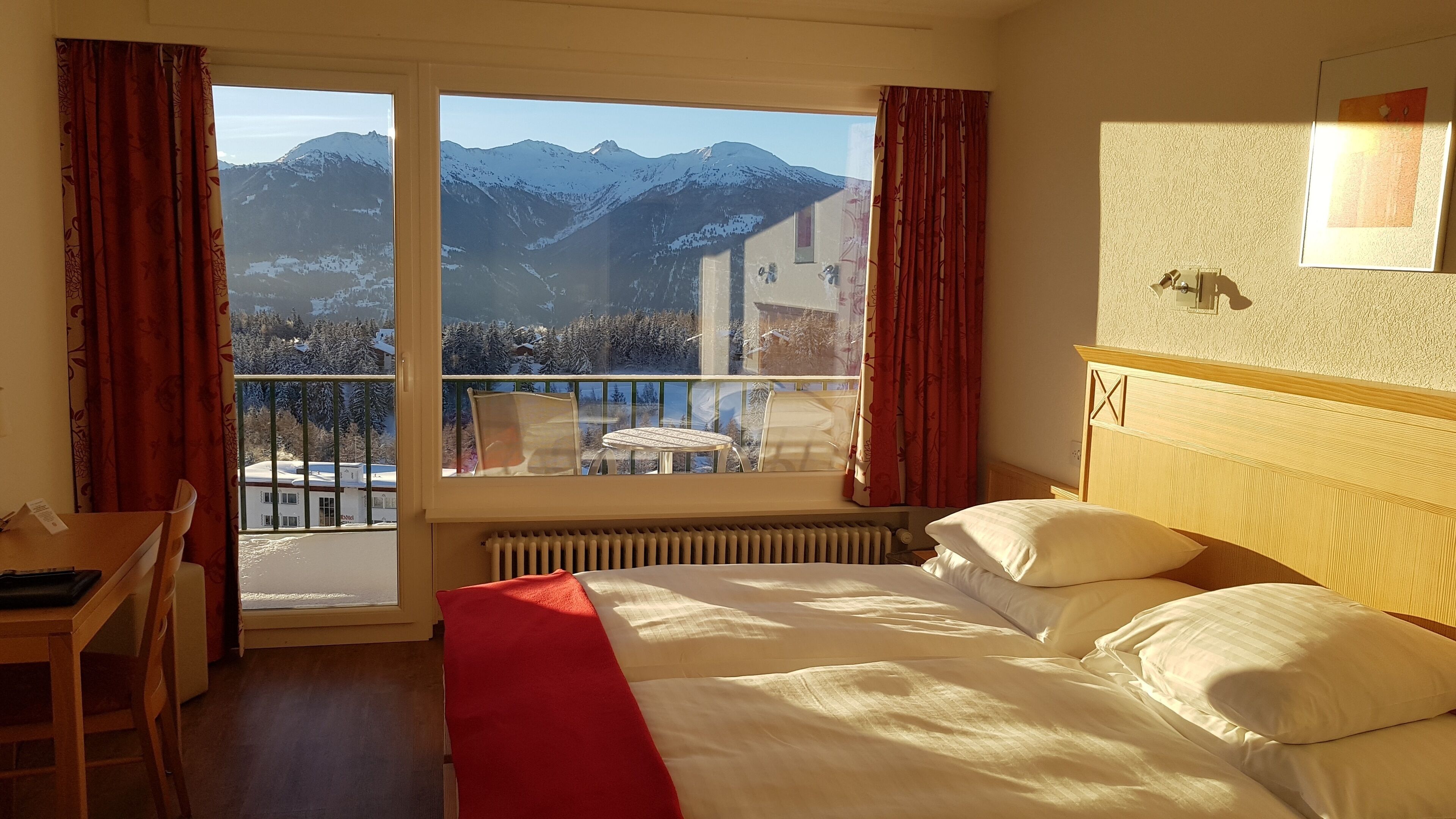 A hotel room with a bed, a desk, and a view of snow-covered mountains through large windows.