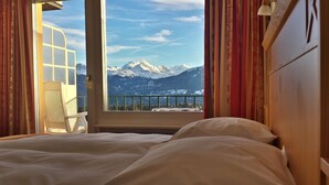 A bedroom with a bed, a window with curtains, and a view of snow-capped mountains. - Hôtel Splendide (Crans-Montana)
