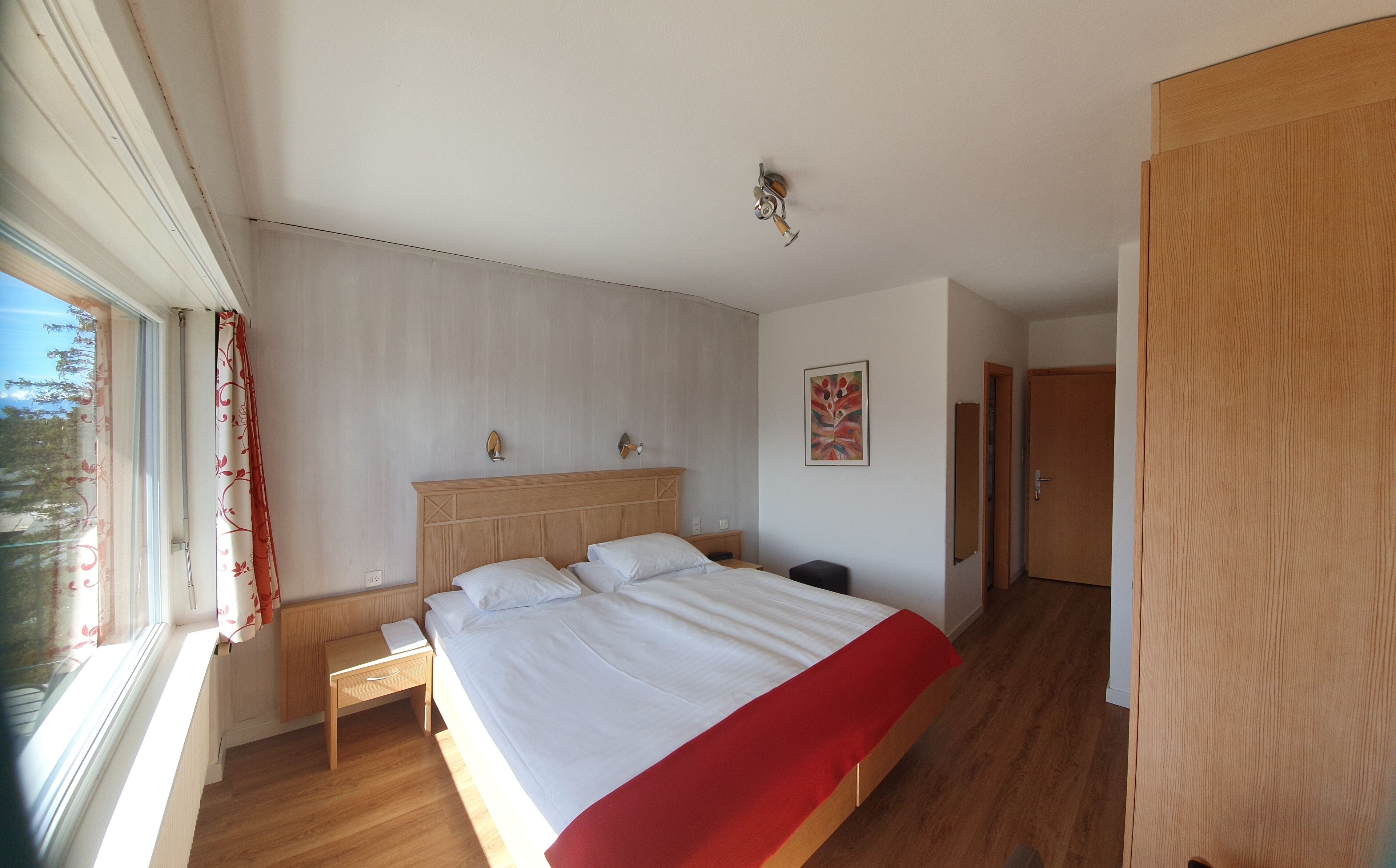 Un dormitorio con una cama de madera, una mesita de noche, una ventana con cortinas, una lámpara de pared y una puerta.