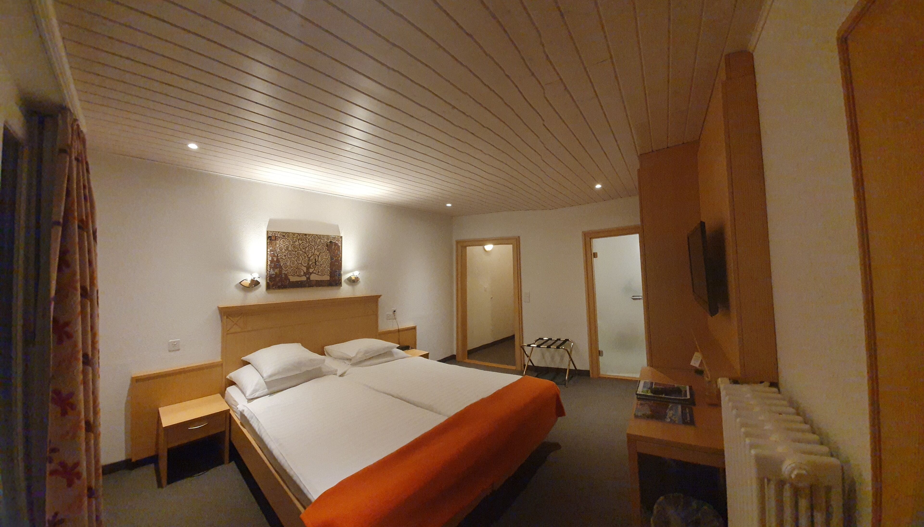 Ein Hotelzimmer mit Holzboden, einem Bett mit weißen Leinen, einem Nachttisch, einem an der Wand befestigten Fernseher und einer Heizung.