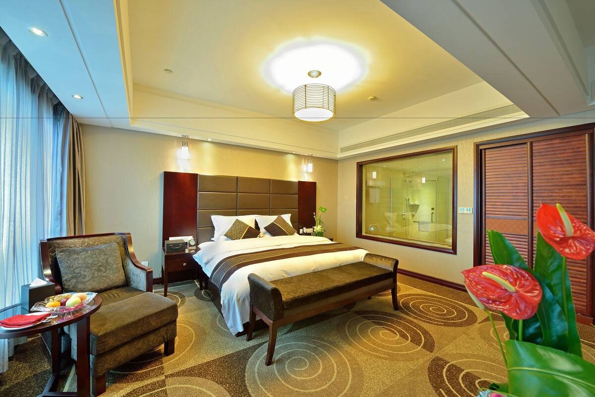 Habitación de hotel con cama, una silla, una mesita con una bandeja de fruta, un banco y un ventanal con persianas.