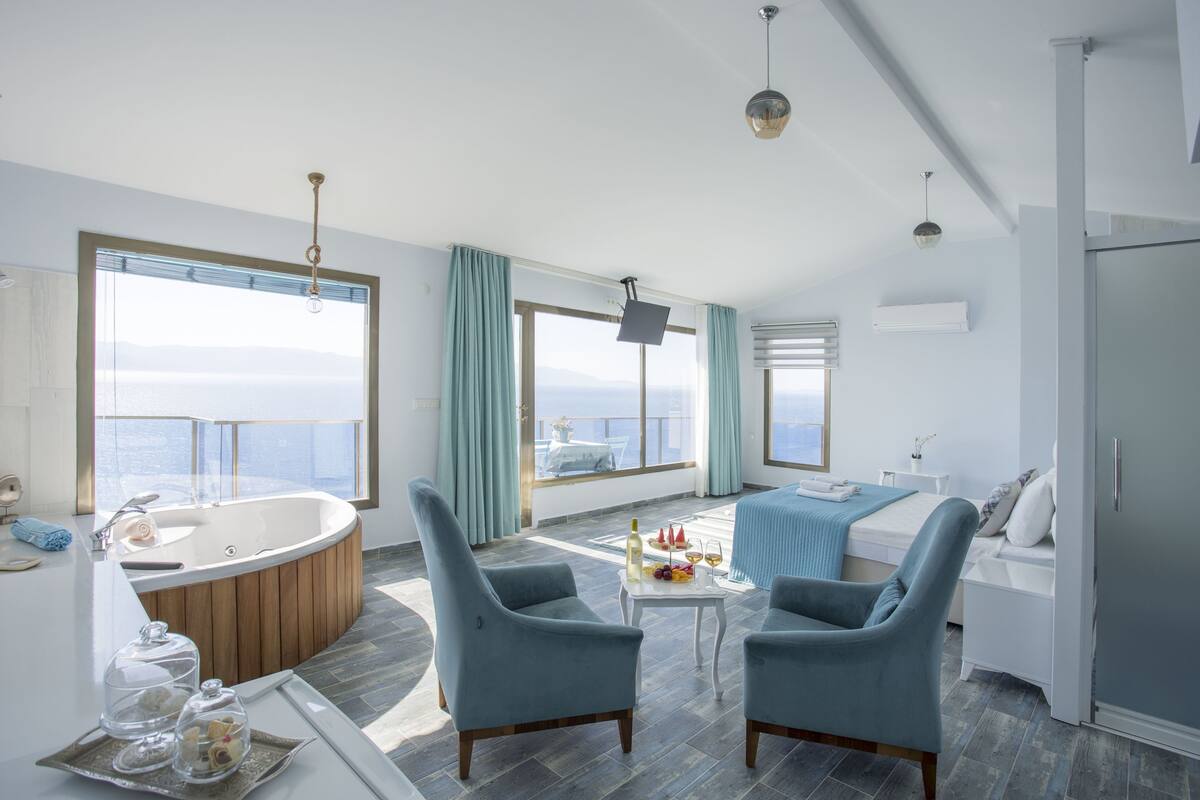A modern hotel room with a large bathtub, two blue armchairs, a small table with a bottle and fruit, and a bed with a blue blanket.