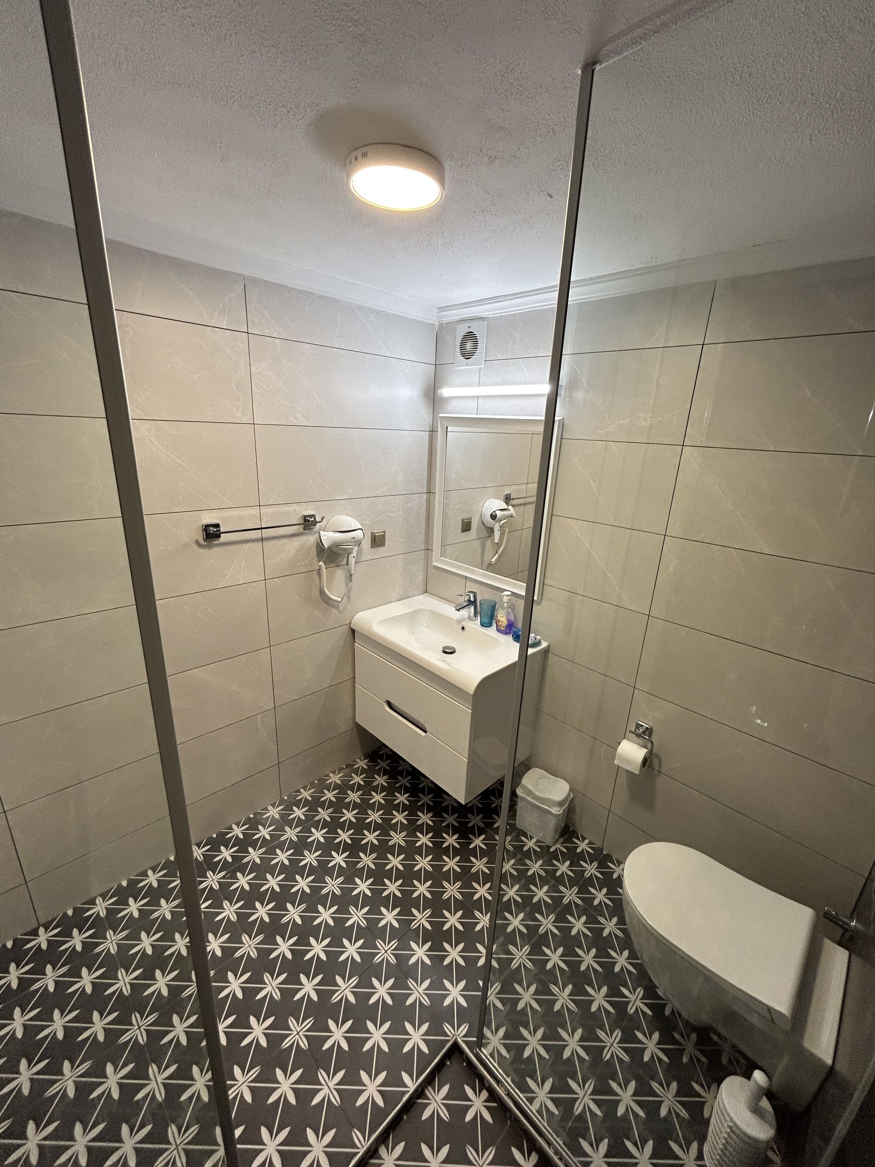 Un baño con lavamanos, espejo y retrete blancos. El piso presenta un estampado de baldosas blanco y negro.