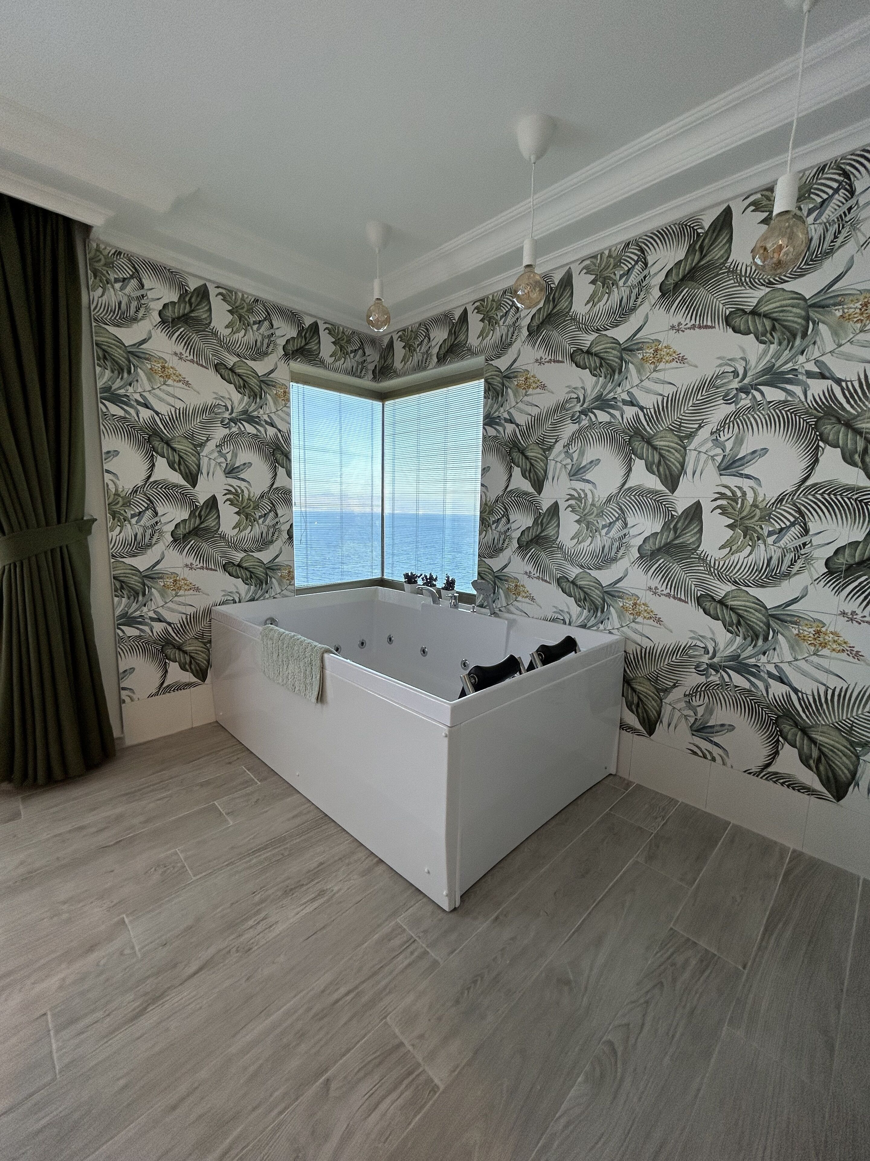 Un baño moderno con una bañera independiente, un ventanal con persianas y papel tapiz con motivos de hojas tropicales.