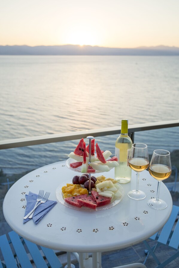 Un tavolo con un vassoio di frutta, una bottiglia di vino e bicchieri, su un balcone con vista sul mare.