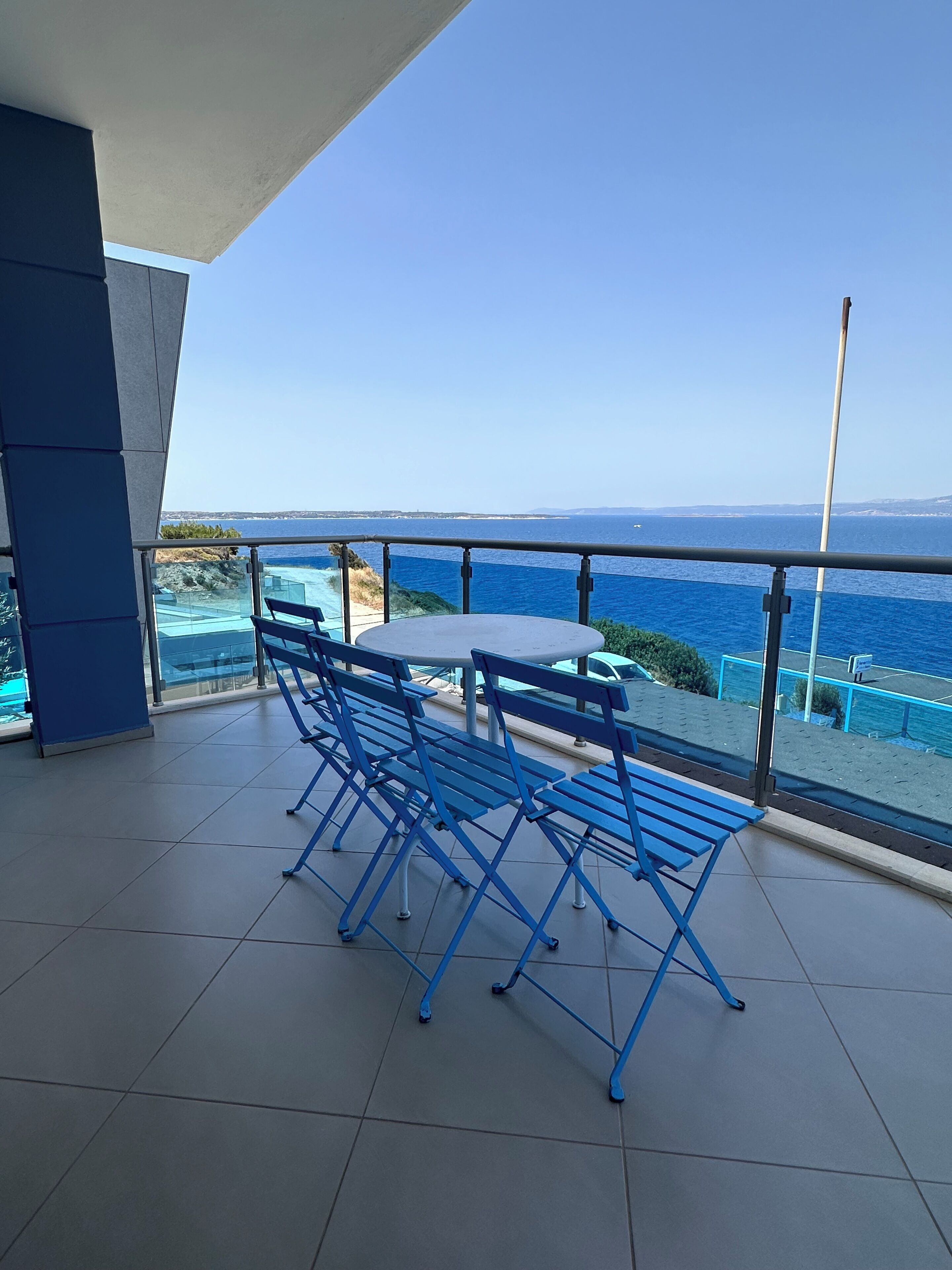 A balcony with blue chairs and a table, overlooking a clear blue sea and distant land.