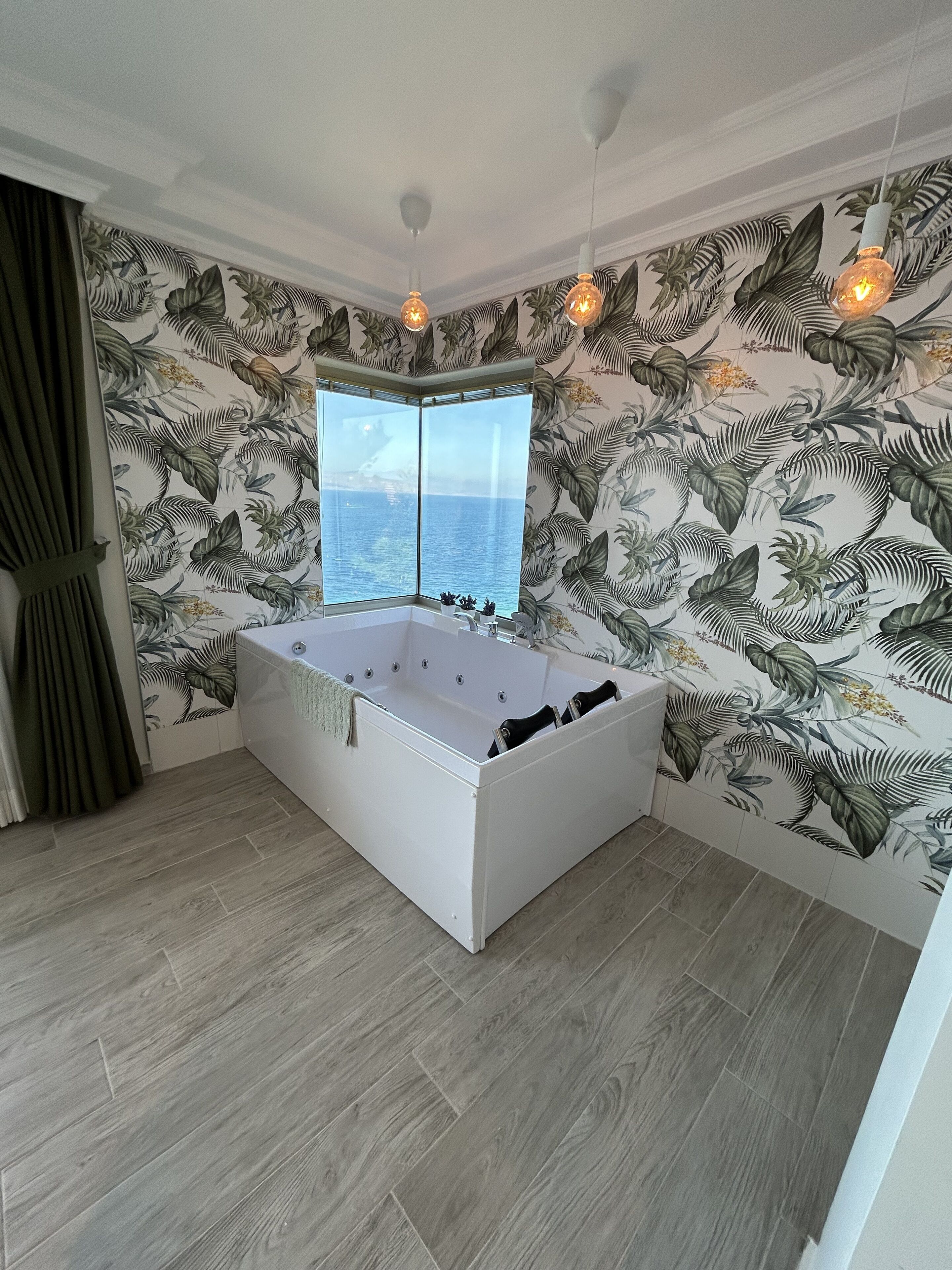 A modern bathroom with a large bathtub, a view of the sea, and tropical wallpaper.