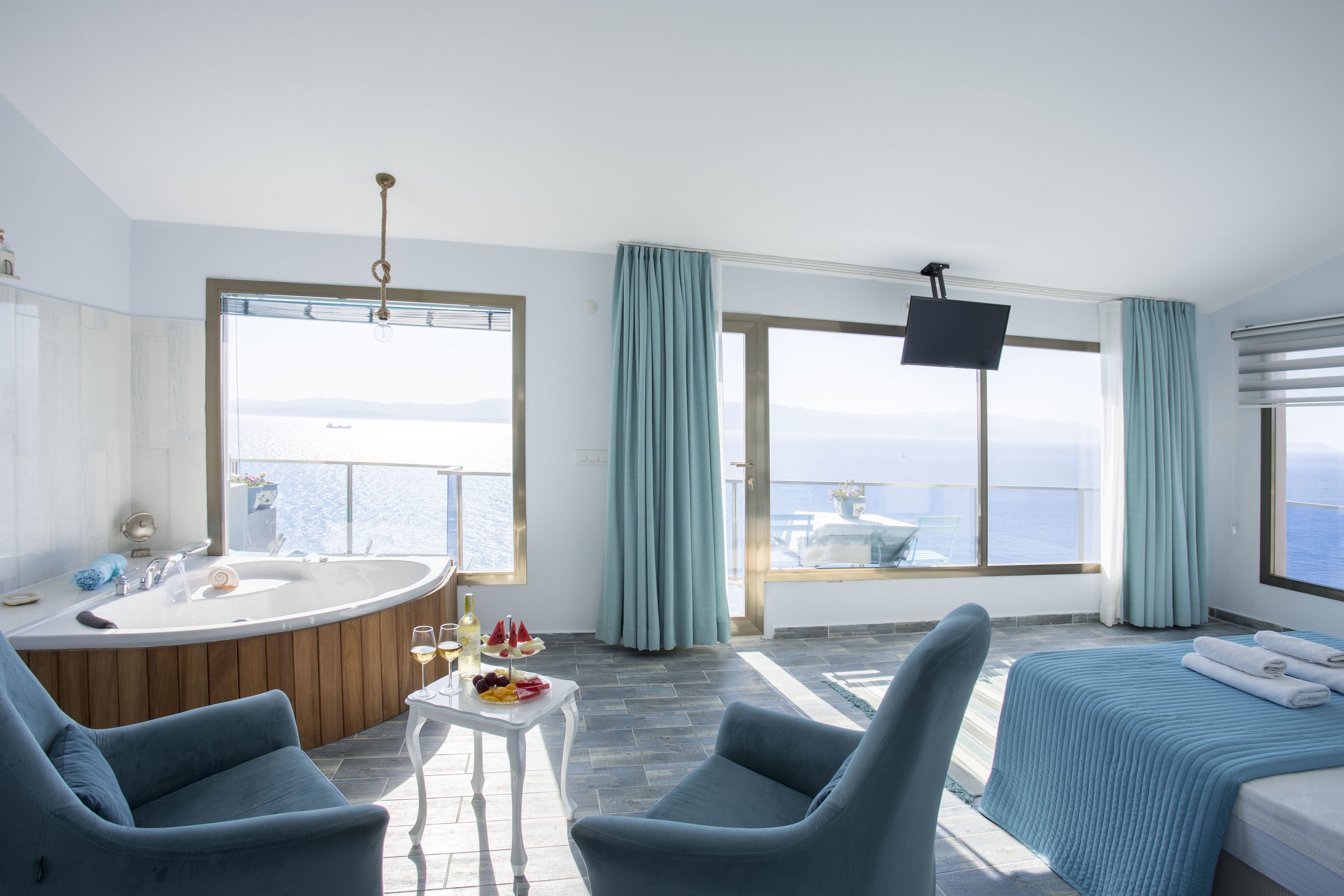 A modern hotel room with a large bathtub, a bed with a blue bedspread, two blue armchairs, a small table with a fruit bowl and glasses, and a large window with a view of the sea.