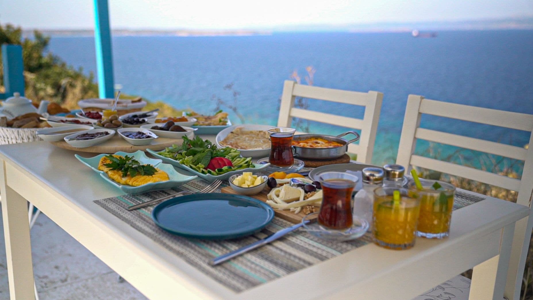 A table set with various dishes and drinks, overlooking a scenic view of the sea.