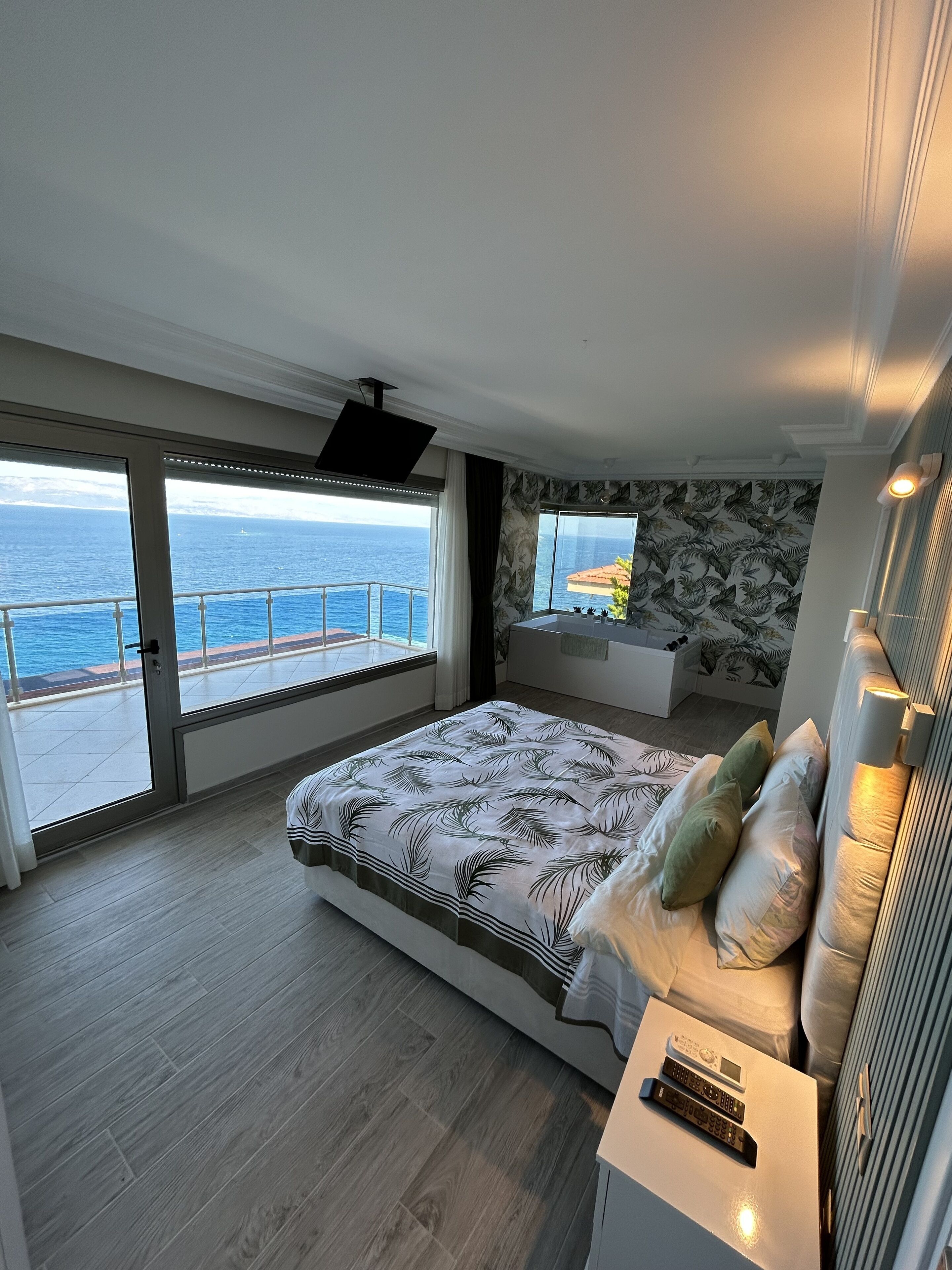 A bedroom with a large bed, a TV, a balcony with a view of the sea, and a window with a leaf-patterned wall.