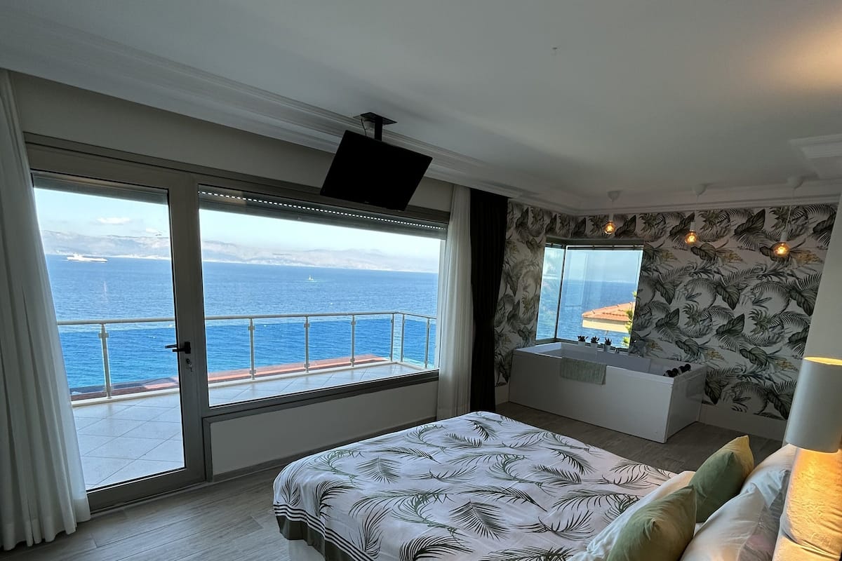 A bedroom with a large bed, a nightstand, a TV, and a view of the sea.