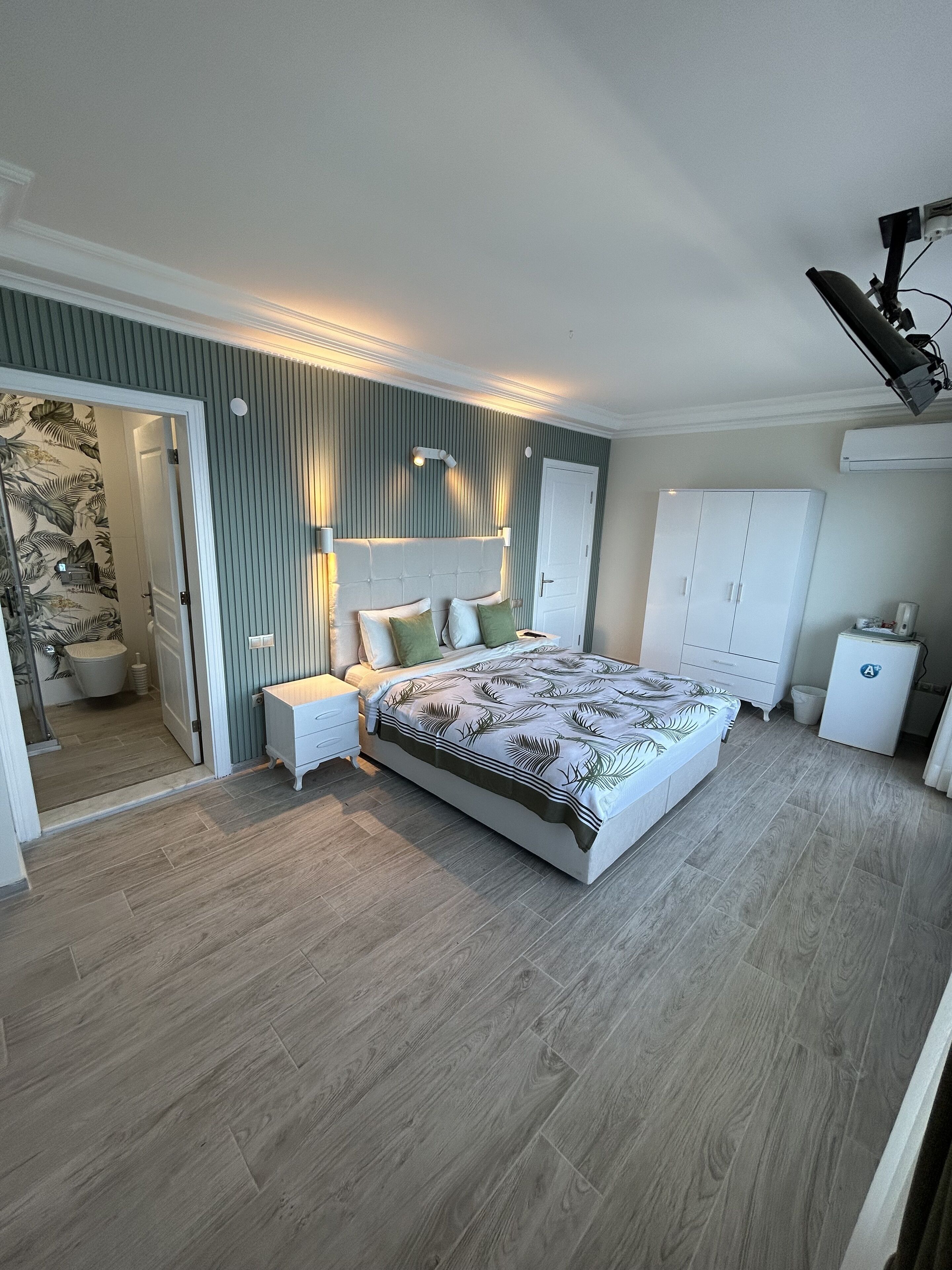 Un dormitorio con una cama grande, un armario, un refrigerador pequeño y un baño visible a través de una puerta abierta.