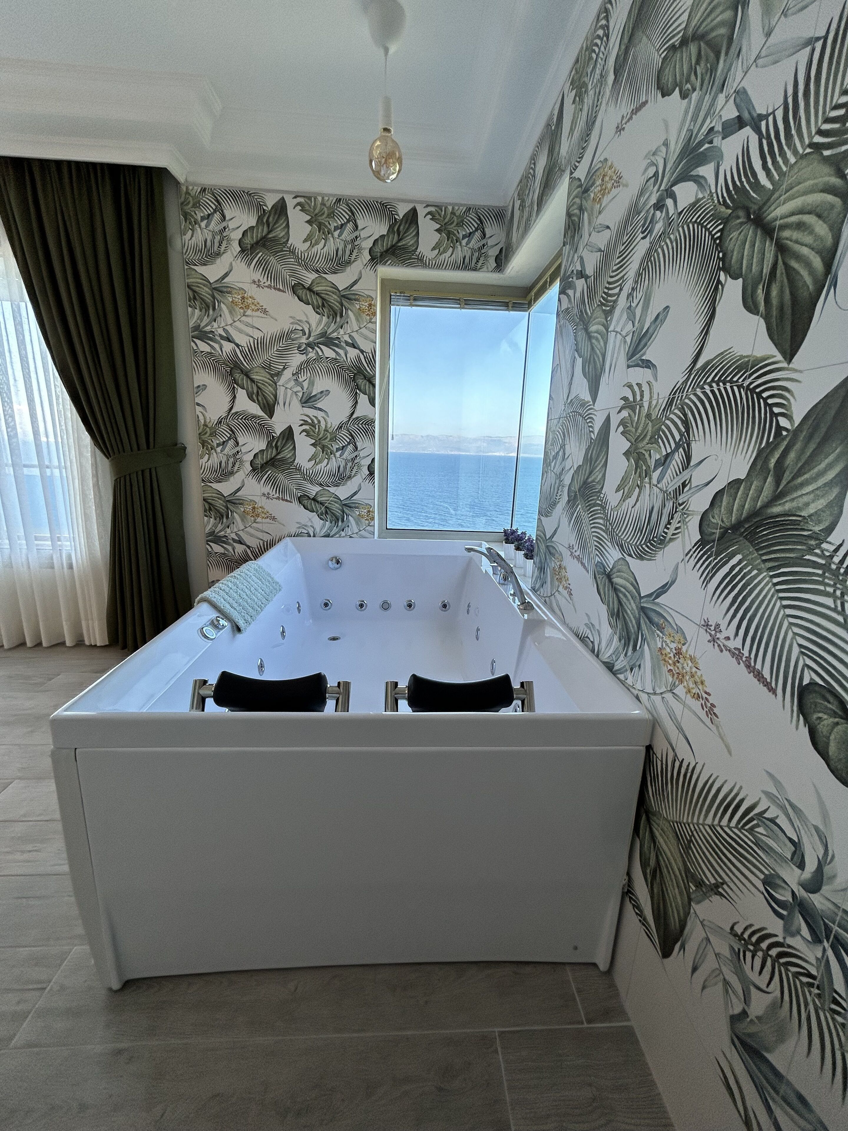 A modern bathroom with a freestanding bathtub, a large window with a view of the sea, and tropical wallpaper.