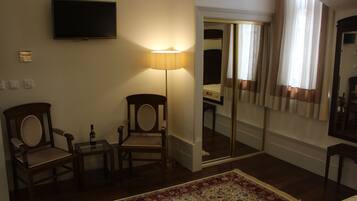 A hotel room with a flat-screen TV mounted on the wall, a standing lamp, two wooden chairs, a small table with a bottle, a patterned rug, and a large mirror reflecting the room.