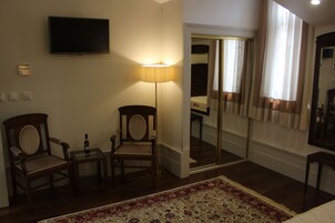 A hotel room with a flat-screen TV mounted on the wall, a standing lamp, two wooden chairs, a small table with a bottle, a patterned rug, and a large mirror reflecting the room.