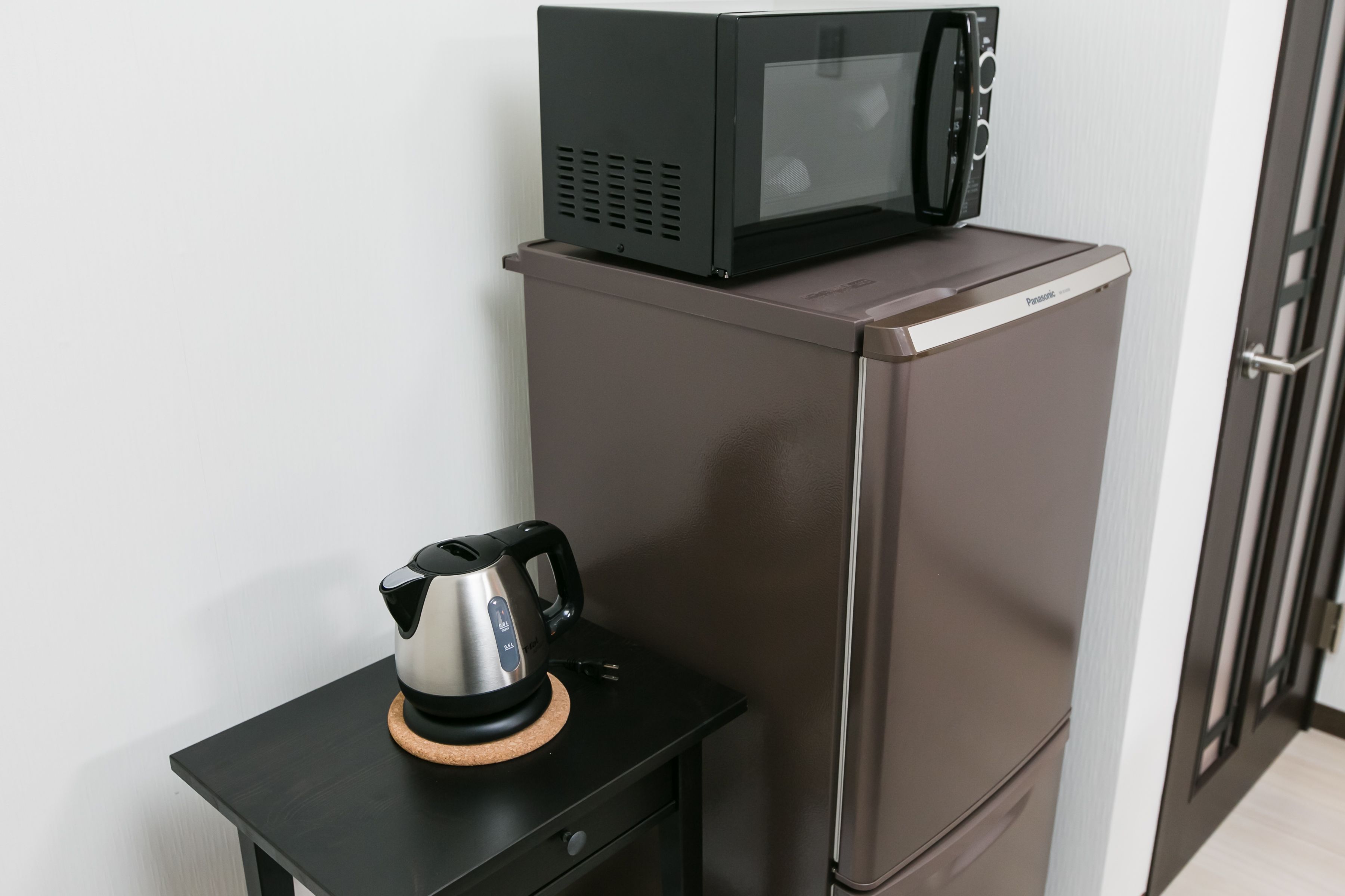 Une petite cuisine équipée comprenant un micro-ondes posé sur le dessus d’un réfrigérateur, une bouilloire sur une petite table et une porte dotée d’une poignée.