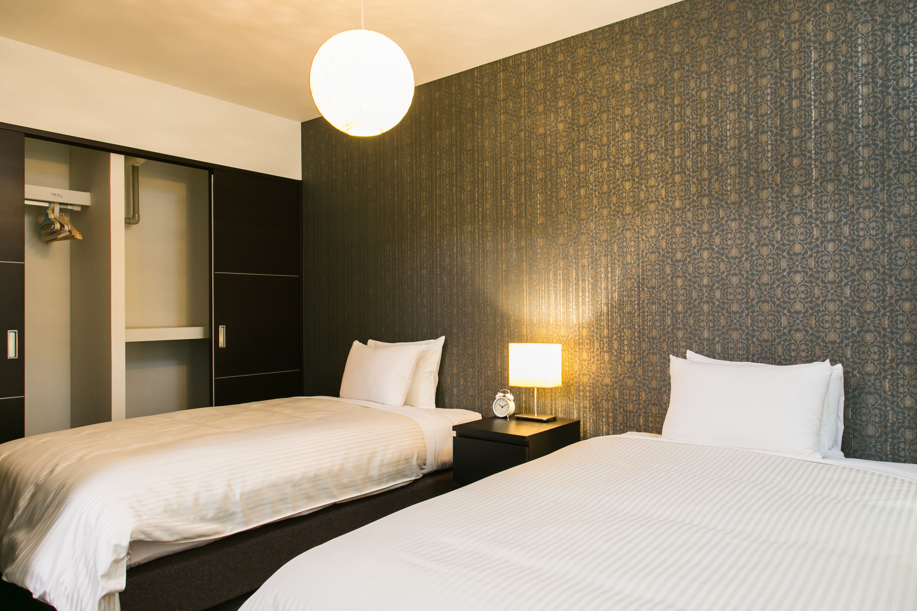 A hotel room with two beds, a wardrobe, a nightstand with a lamp, and a patterned wall.