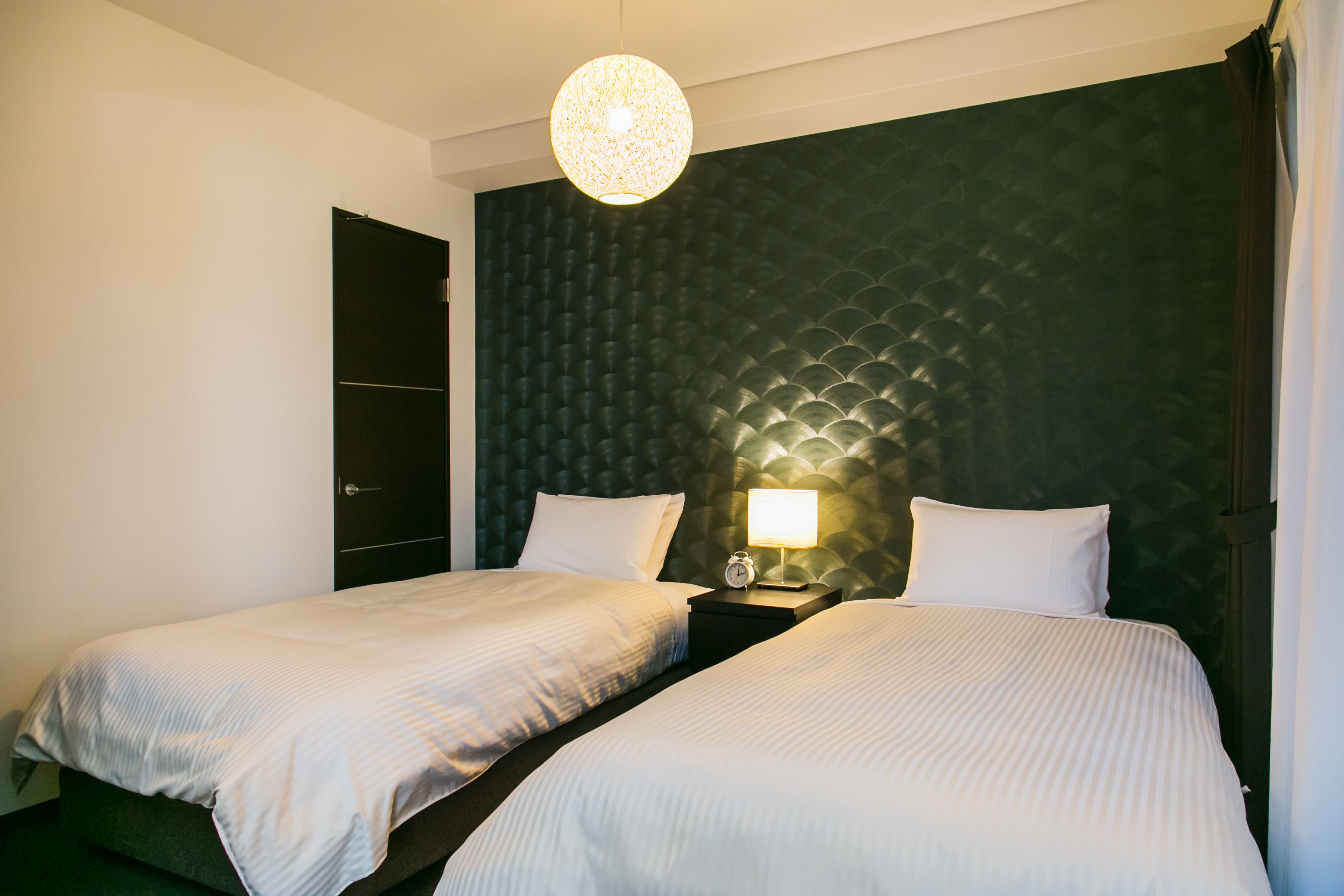 A hotel room with two beds, a dark green headboard, a round light fixture, and a nightstand with a lamp.