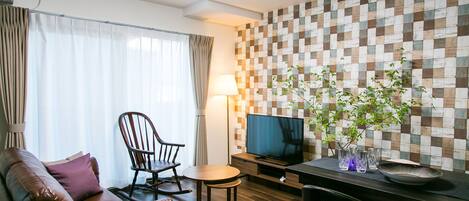 A modern living room with a leather sofa, a wooden rocking chair, a small round table, a flat-screen TV, and a decorative wall with a checkered pattern.