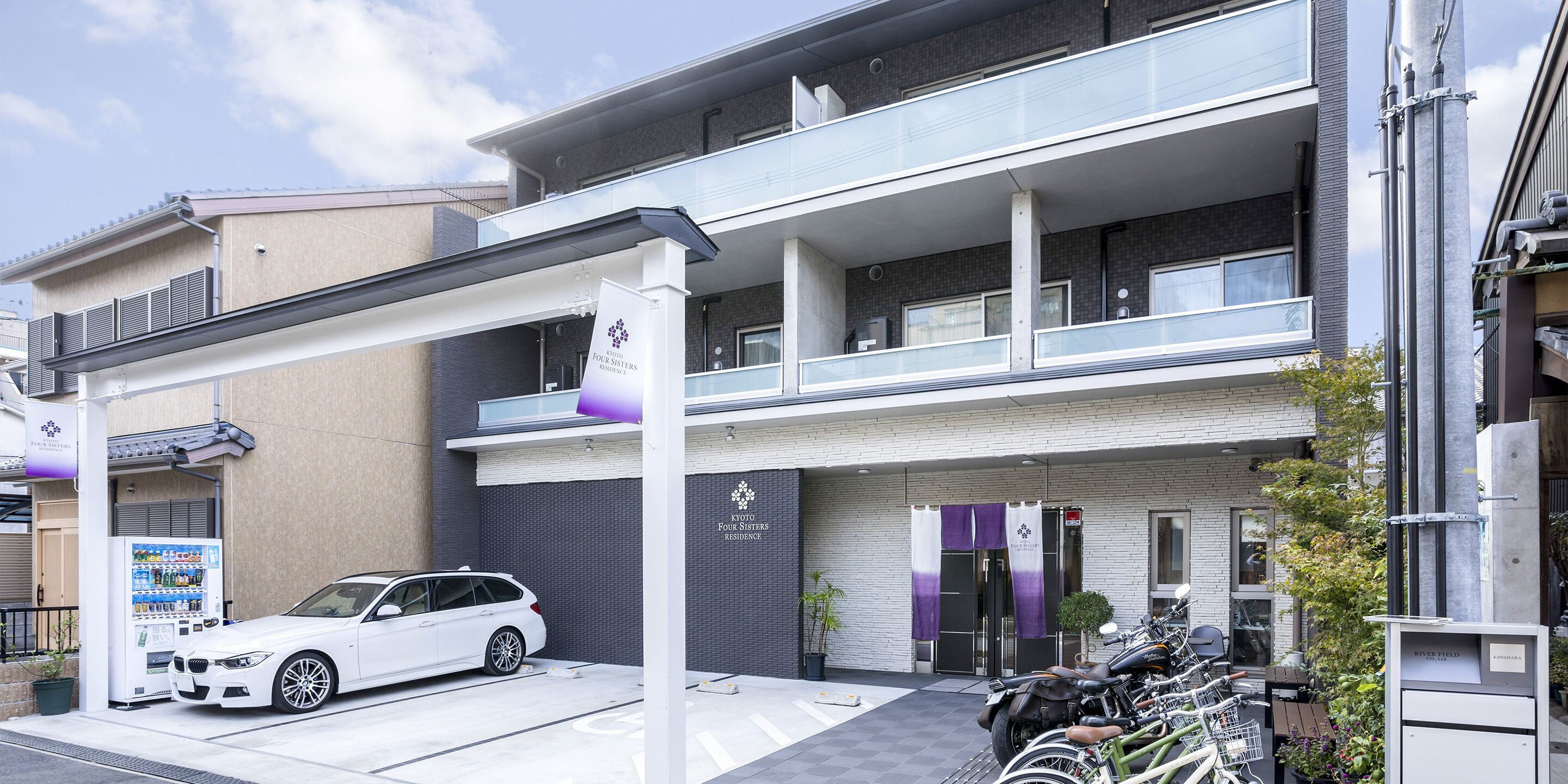 A modern building with a car parked in front, a vending machine, and bicycles parked outside.