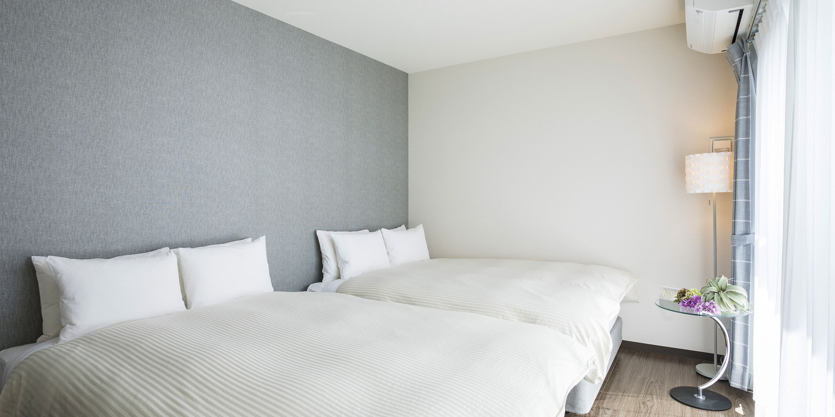 Een hotelkamer met twee bedden, een grijze structuurwand, een klein nachtkastje met een lamp en een raam met gordijnen.