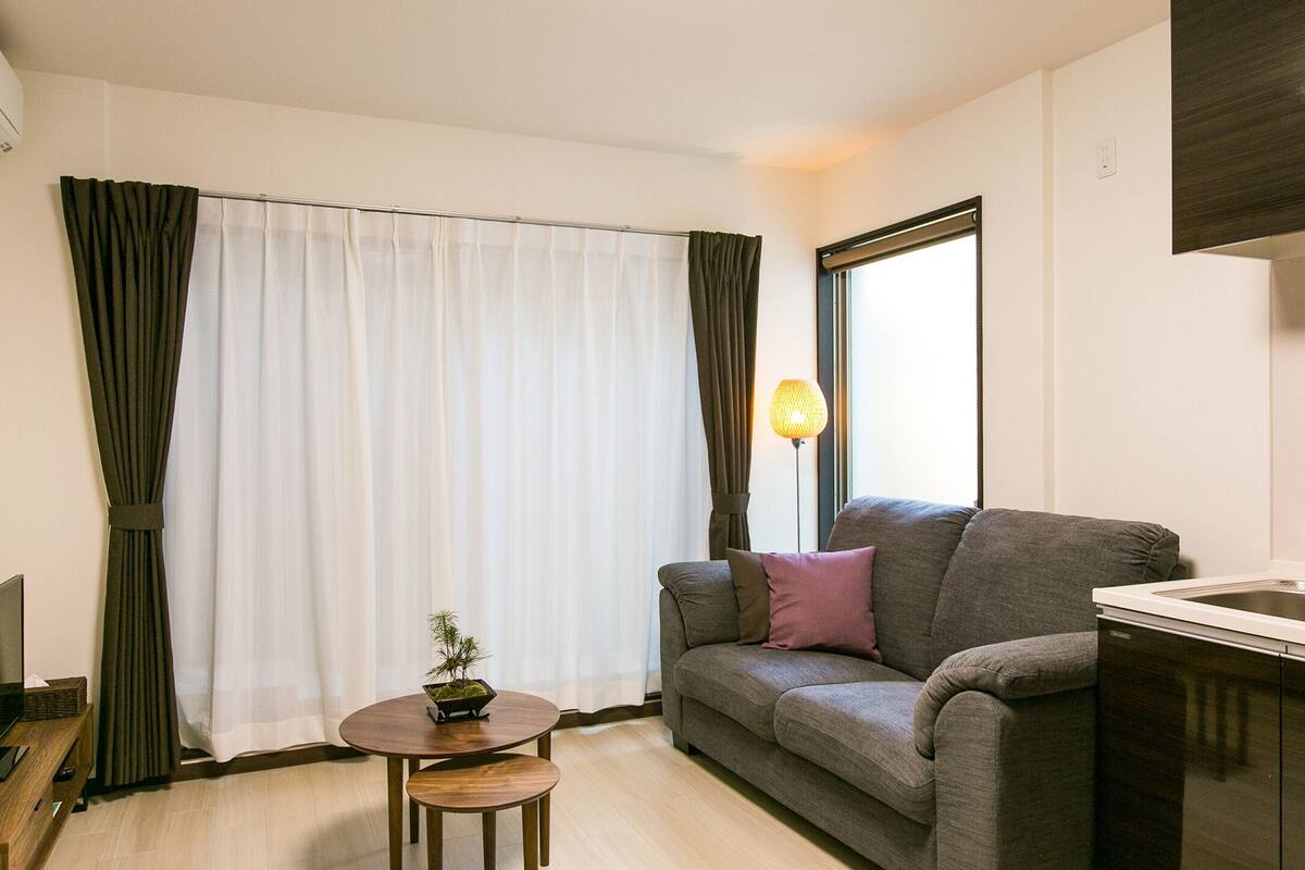 Ein modernes Wohnzimmer mit einem grauen Sofa, einem kleinen runden Tisch mit einer Pflanze, einem Fernseher auf einem Holzschrank, einem Fenster mit Vorhängen und einer Küchenzeile mit Spüle und Schränken.