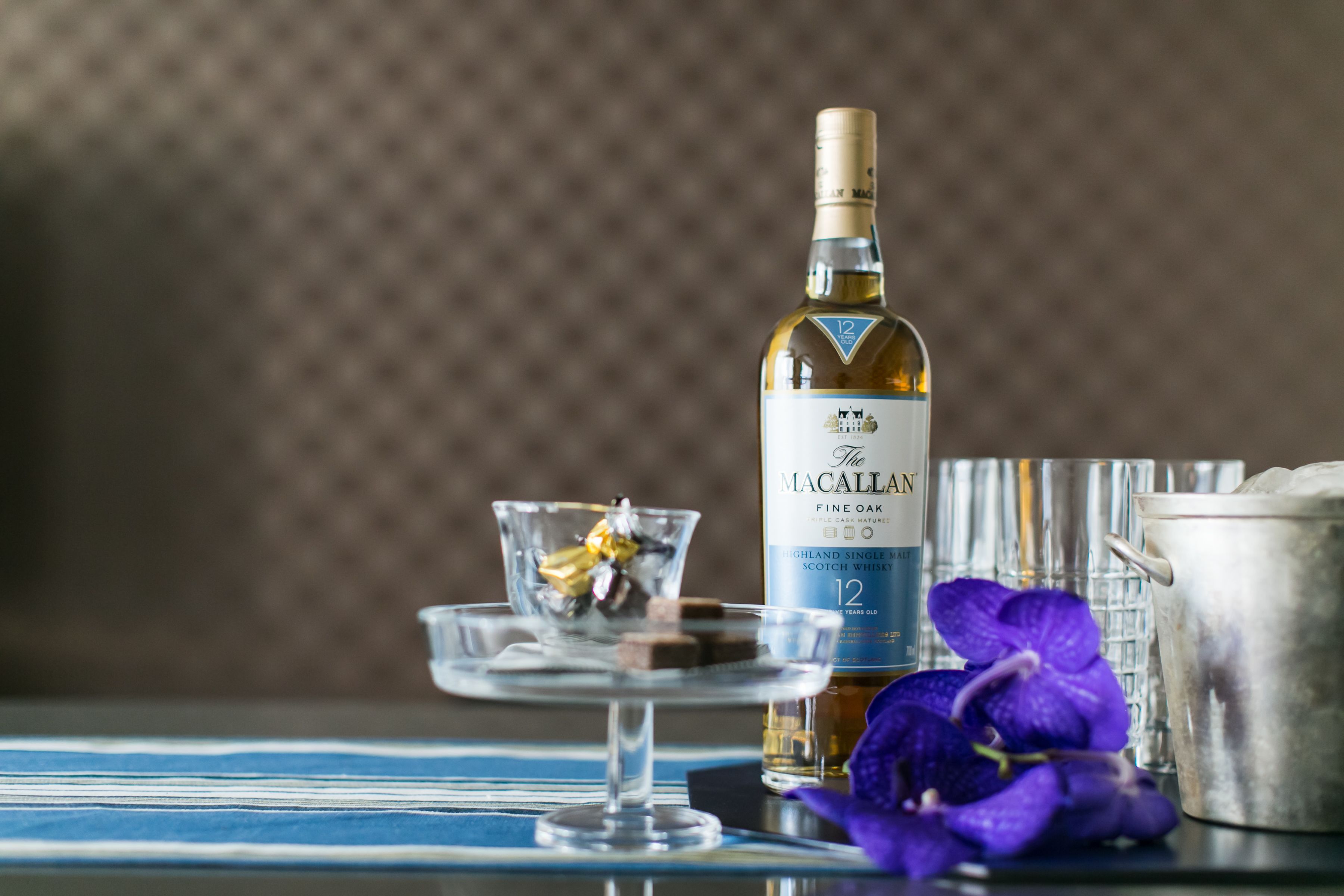 Une bouteille de whisky The Macallan, un verre orné d’une fleur et un récipient en métal sur une table.