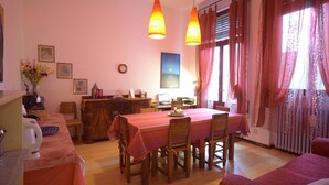 Un comedor con una mesa preparada para cuatro, sillas de madera, una cafetera y un florero con flores en la encimera.