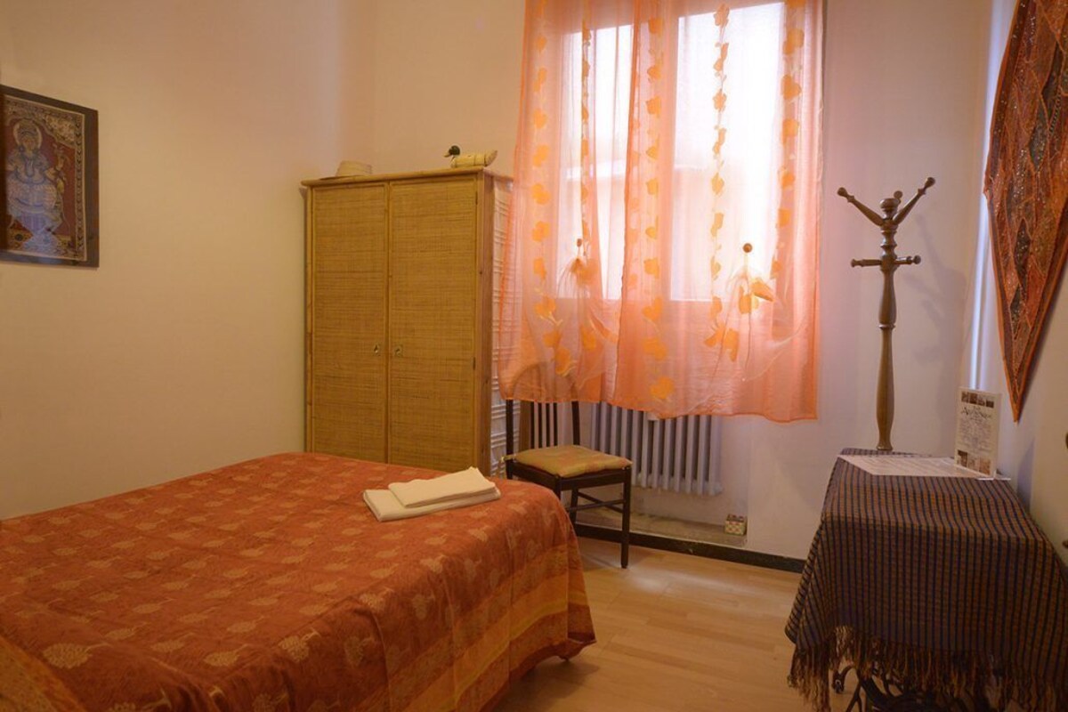 A bedroom with a bed, a wooden wardrobe, a small table with a cloth, a chair, and a radiator.
