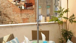 Una mesa redonda con sillas de mimbre y un tablero de vidrio, ubicada en un balcón con vistas a edificios y una planta.