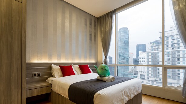 A modern hotel room with a large bed, a nightstand, and a view of the cityscape through a large window. - Soho Suites KLCC By Aloha (Kuala Lumpur)