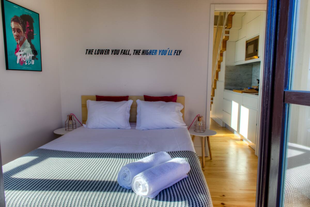 A hotel room with a bed, a wall with a motivational quote, a small table with a candle, and a view of the kitchen through an open door.