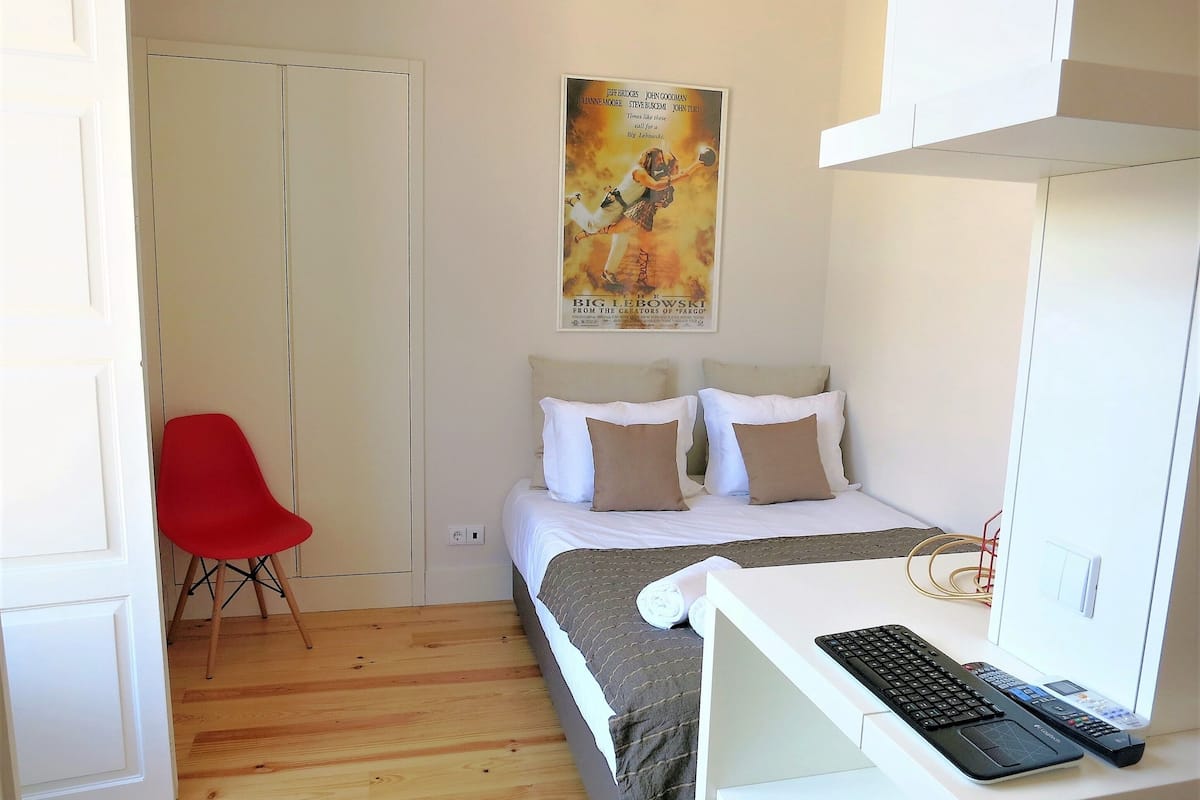 A small, neatly arranged bedroom with a single bed, a desk with a computer, and a red chair.