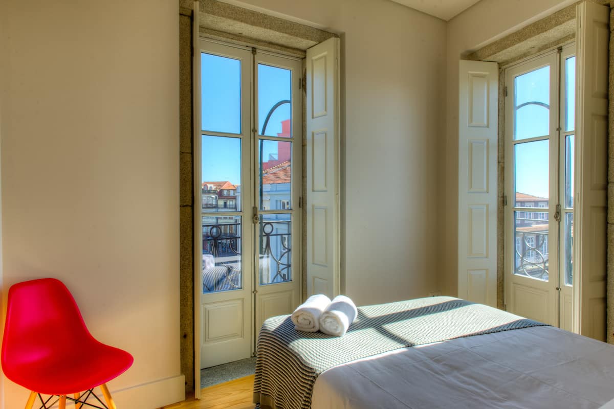 A hotel room with a bed, a red chair, and large windows with a view of buildings outside.