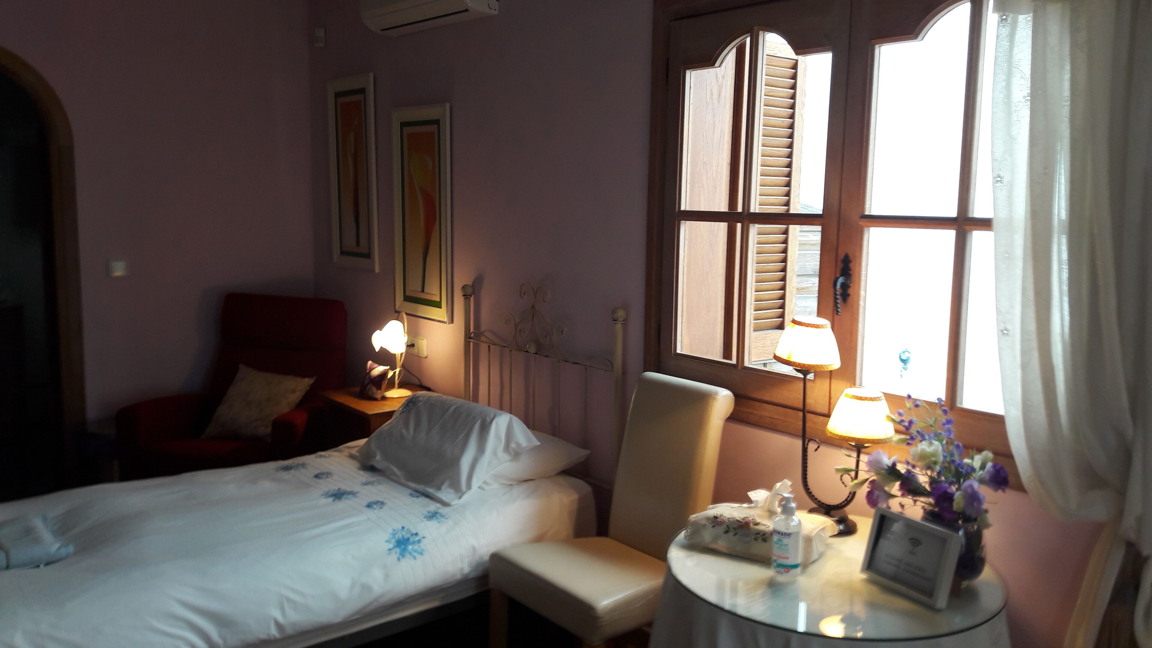 A hotel room with a bed, a chair, a small table with a lamp, and a window with shutters.