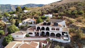 A large, white villa with multiple arches and a tiled roof, surrounded by lush greenery and hills.